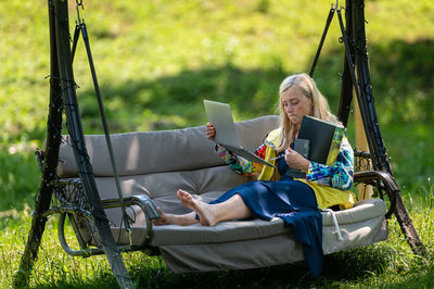 Mature woman with laptop and documents working in garden on rocking couch, green home office concept