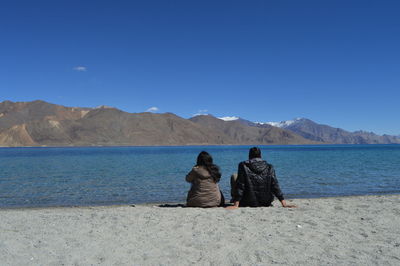 Rear view of man and woman sitting at lakeshore against blue sky on sunny day