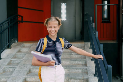 A smiling teenage girl with a backpack and a folder in her hands goes down the stairs