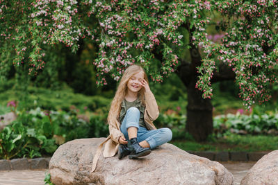Happy girl child with long wavy brown hair sits on a stone in spring in a blooming park and smiles
