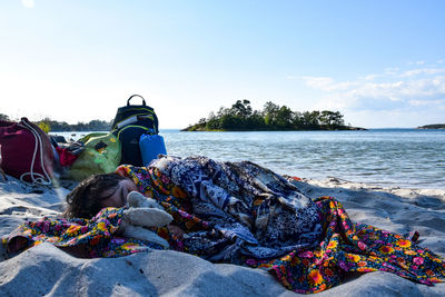 Boy under blanket relaxing at sandy beach against sky