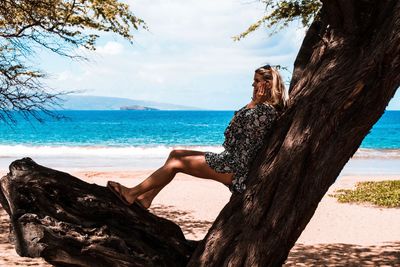 Side view of young woman leaning on tree trunk at beach