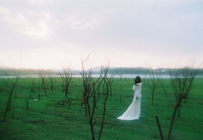 Woman wearing white dress while standing on grass against sky during sunset