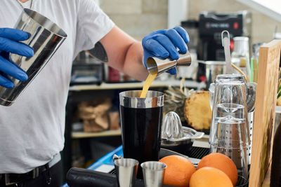 Crop anonymous male barkeeper in casual clothes and gloves pouring fresh citrus juice into shaker while preparing mixed beverage in cafe