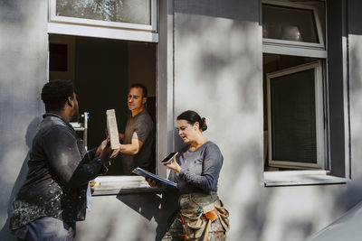 Workers passing plank through window while female construction manager talking on phone outside house