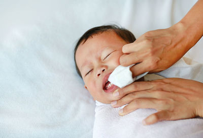 Cropped hand of mother cleaning baby boy teeth lying on bed at home