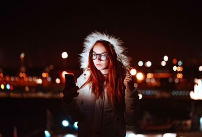 Woman in warm clothes using mobile phone while standing outdoors at night