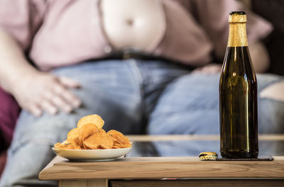 Midsection of senior man with beer and potato chips on table sitting at home