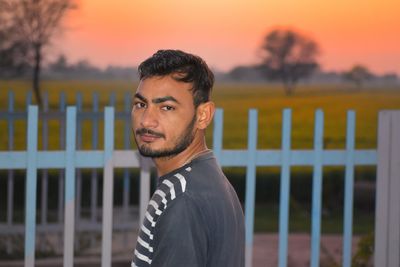 Portrait of young man standing against fence during sunset