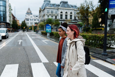 Side view of cheerful stylish multiethnic girlfriends walking and crossing marked road in city in gray day