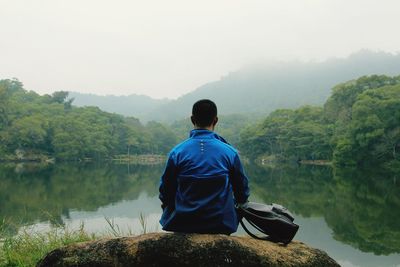 Rear view of man sitting on rock by lake against sky