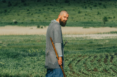Bald bearded man in metal chain mail over linen shirt stands in middle of field, holding sword.