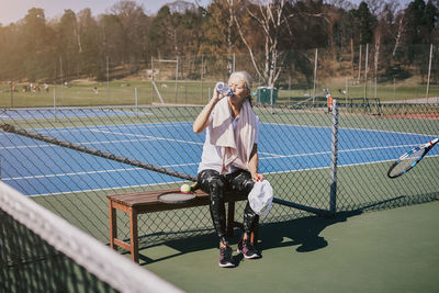 Tired senior woman drinking water while sitting on bench at tennis court