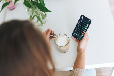Young woman checking cryptocurrency charts and stock quotes on mobile phone with cup of coffee 