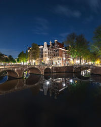 Bridge over river by illuminated buildings against sky at night