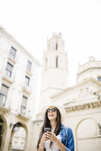 Low angle view of cheerful young woman using smart phone against building