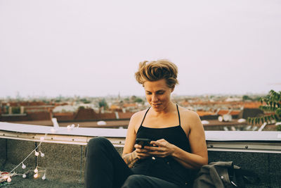 Mid adult smiling woman using mobile phone while sitting on terrace against sky in city