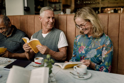 Happy senior man and woman talking with each other while reading books by male friend in cafe