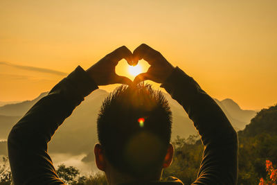 Rear view of man forming heart shape with hands against sky during sunset