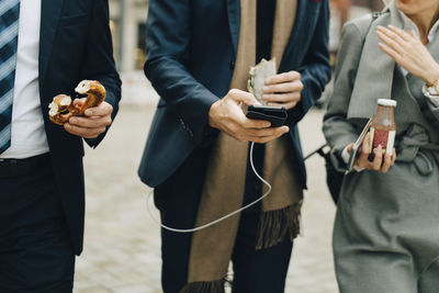 Midsection of business professionals holding pretzel and drink while using smart phone in city