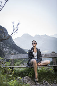 Full length of young woman sitting on bench against mountains