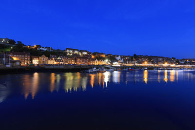 Illuminated buildings by river against clear blue sky at night