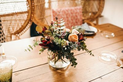 From above bouquet of miscellaneous flowers and green plant twigs in vase with water on a wooden table set for a meal with beautiful designed rattan chair on the background