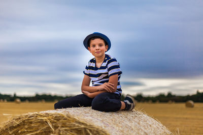 Portrait of cute boy sitting on hay bale against cloudy sky