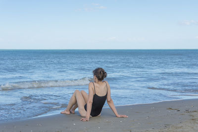 Rear view of woman sitting on shore at beach against sky