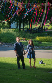 Full length of couple looking at ribbons decorated on branch at public park