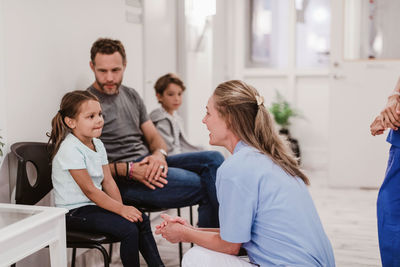 Female pediatrician talking with girl sitting by family in hospital corridor