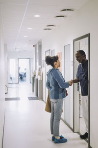 Full length of smiling doctor greeting female patient standing in corridor at hospital