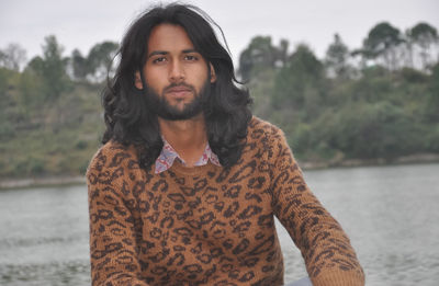 A attractive indian young guy with long hair and beard looking at camera while sitting by the lake