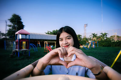 Portrait of young woman making heart shape against sky at playground