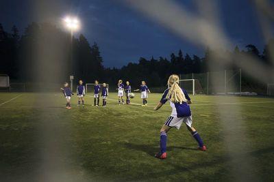 Girls playing soccer seen from goal post net on field against sky