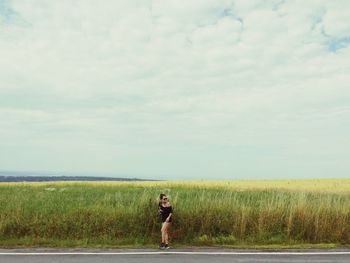 Woman standing on grassy field by road against cloudy sky