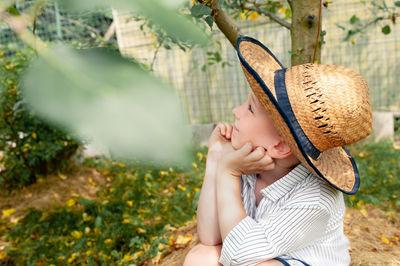 Cute pre-school boy in a straw hat and a rustic white shirt looks at an apple tree in home garden