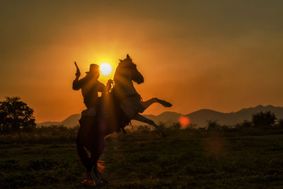 Silhouette person holding gun while horseback riding on land against sky during sunset