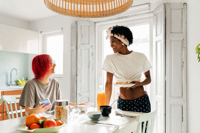 Delighted young lady with bright pink hair smiling and browsing smartphone while sitting at table near cooking african american friend during breakfast at home