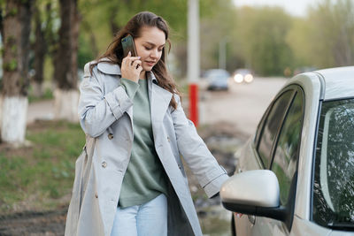 Beautiful girl with long hair in a grey trench coat using smartphone call gets into the car