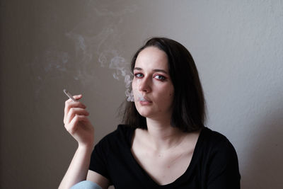 Portrait of young woman with cigarette sitting against wall at home