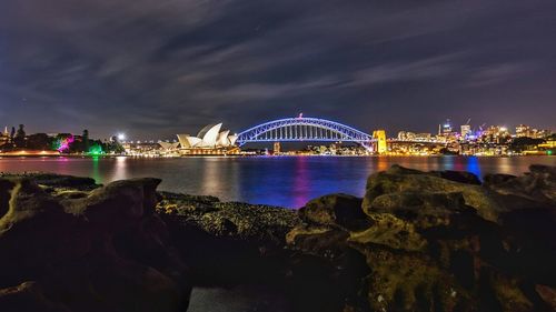 Illuminated city at waterfront against cloudy sky