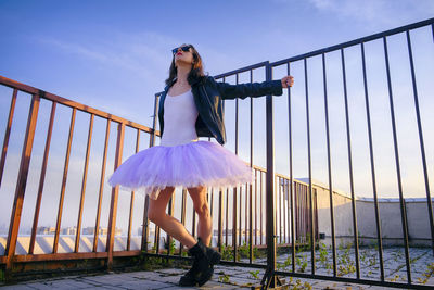 A ballerina in tutu stands at the fence on the roof at sunset in boots and a jacket