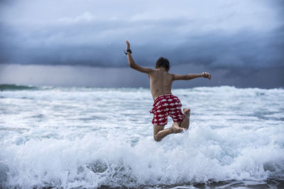 Rear view of shirtless boy jumping in sea against cloudy sky