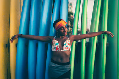 Young african american female wearing colorful bikini top and headband spreading arms while standing back to colorful surf boards