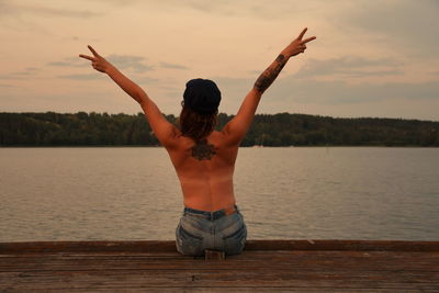 Rear view of shirtless woman gesturing while sitting on pier over lake against sky during sunset