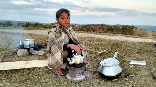 Portrait of girl with short hair preparing food while crouching on field against sky during sunset