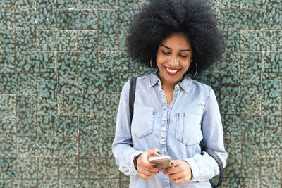 Smiling young woman using phone while standing against wall