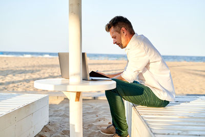 Side view of focused male entrepreneur in smart casual clothes sitting on lounger and writing in notebook while working on remote project on sandy beach near sea