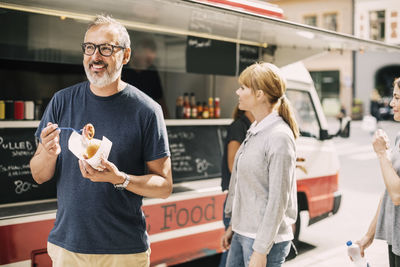 Mature man eating food while standing against truck at street